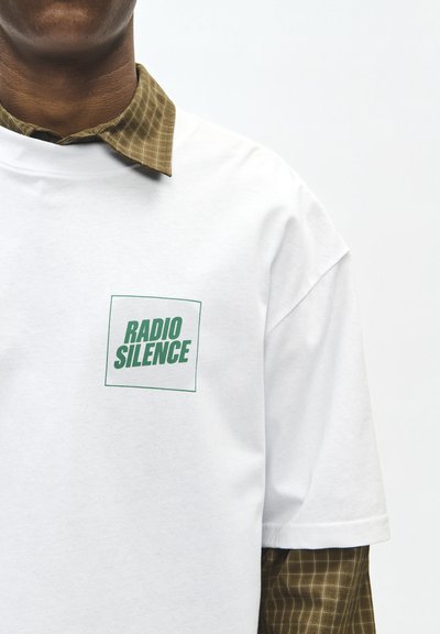 Personne portant un t-shirt blanc avec le texte vert "RADIO SILENCE" sur une chemise à carreaux marron avec col visible.
