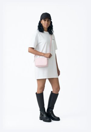 Nainen pukeutuneena valkoiseen ylisuurikokoiseen t-paitamekkoon, mustiin polven korkeisiin saappaisiin ja mustaan lippikseen, kantaen vaaleanpunaista olkalaukkua, seisoo valkoisen taustan edessä.