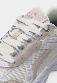 Κοντινό πλάνο άσπρου και μπεζ αθλητικού παπουτσιού με δίχτυ, λευκά κορδόνια και το λογότυπο "GAP" στη γλώσσα και την πλαϊνή πλευρά.