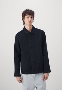 Chaqueta de color azul oscuro, abotonada, con cuello, dos bolsillos frontales y un corte relajado. Fabricada con una tela resistente, presenta un diseño minimalista.