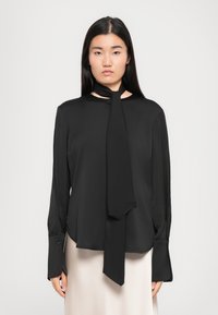 Ženska v dolgem črnem bluzu z podaljšanimi manšetami in pritrjenim metuljčkom, kombinira z svetlo bež krilom.