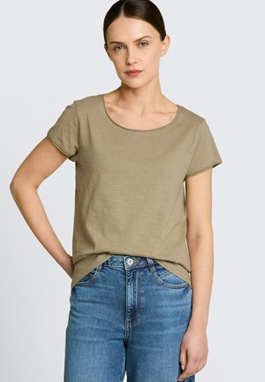 Woman with dark hair tied back wearing a short-sleeve olive green t-shirt and blue jeans, standing with one hand on her hip.