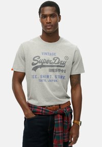 Grå bomulds t-shirt med et vintage print i mørkeblå og lilla. Modellen bærer mørke jeans og en rødternet skjorte bundet rundt om livet.