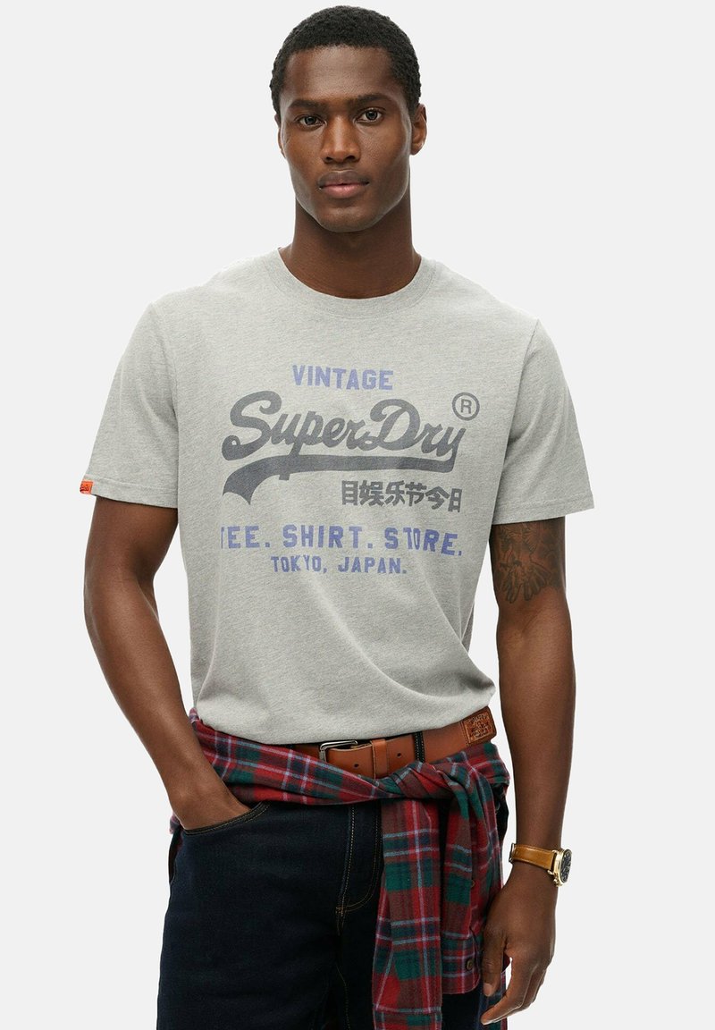 Grå bomulds t-shirt med et vintage print i mørkeblå og lilla. Modellen bærer mørke jeans og en rødternet skjorte bundet rundt om livet.