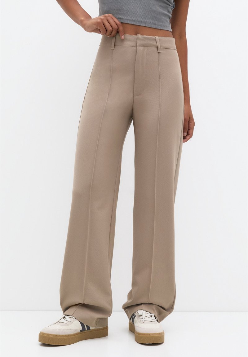 Pantalon large beige à texture lisse, taille haute, plis avant et coupe ajustée, associé à des baskets blanches.