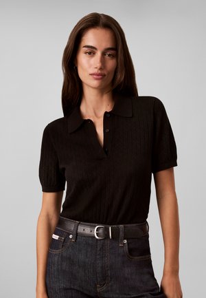 Femme aux longs cheveux bruns portant une chemise noire à manches courtes avec col, rentrée dans un jean foncé avec une ceinture noire, sur un fond gris.