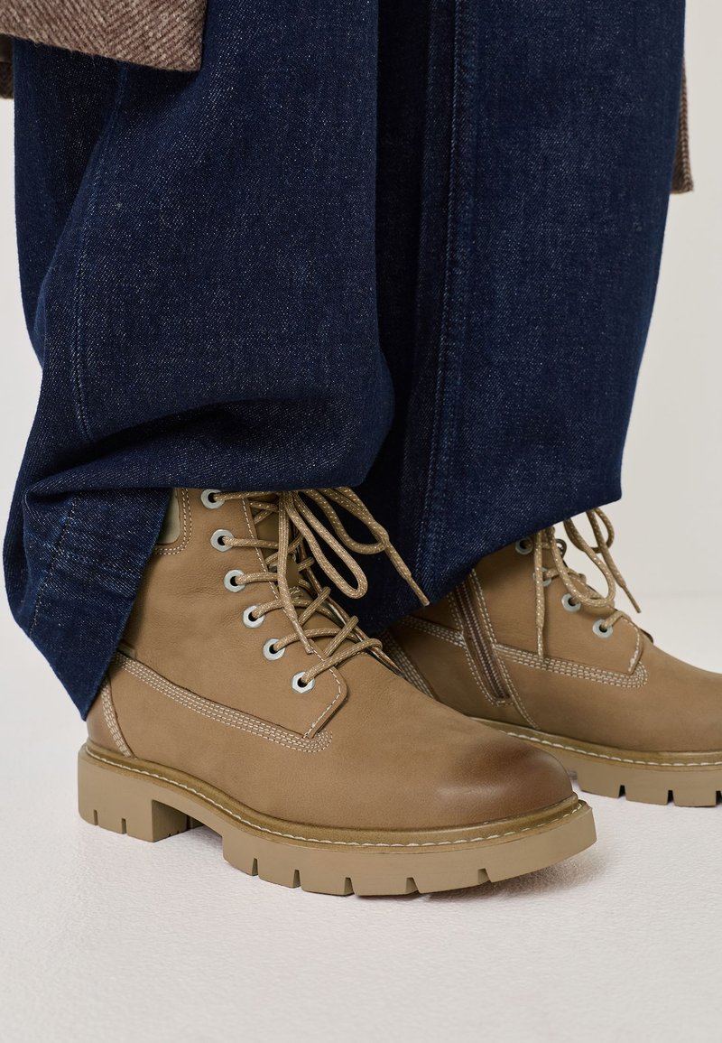 Vaskettomaiset ruskeat nilkkasaappaat, joissa on pyöreä kärki, nauhakiinnitys, metalliset aukot ja teksturoitu kumipohja. Yhdistetty tummiin farkkucapreihin.