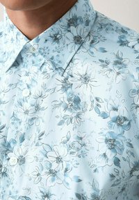 Camicia floreale azzurro chiaro con colletto button-down, caratterizzata da bottoni bianchi e una consistenza morbida. Il design include intricati motivi floreali e foglie.