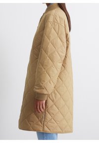 Kobieta ubrana w długi, beżowy płaszcz o wzorze diamentowym z prążkowanymi mankietami, stojąca w bok na jednolitym białym tle.