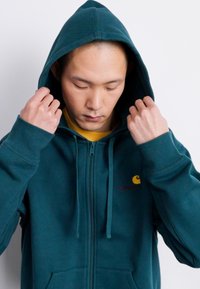 Mėlynos spalvos zip-up bluza su reguliuojamu gobtuvu, raukšlėmis aprašais ir priekinių kišenių. Kairėje krūtinės srityje yra logotipas. Pagaminta iš medvilnės mišinio.