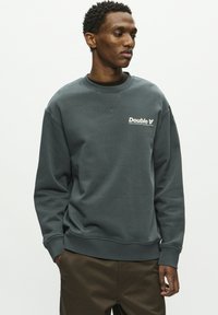 Mørkegrå sweatshirt i et klassisk crew neck design med et hvidt logo på brystet samt ribbede manchetter og kant. Glat tekstur.