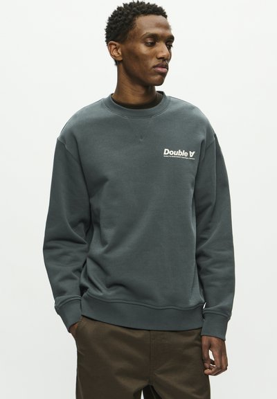 Sweatshirt gris foncé avec un design classique à col rond, arborant un logo blanc sur la poitrine ainsi que des poignets et un ourlet côtelés. Texture lisse.