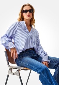 Blauw-wit gestreept overhemd met knopen en gerimpelde manchetten, gecombineerd met een blauwe spijkerbroek. Model draagt zwarte zonnebril en zit op een houten stoel.