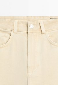 Nahaufnahme des Taillenbunds einer beigen Jeanshose mit Knopf, Gürtelschlaufen, Vordertaschen und sichtbaren Nahtdetails.