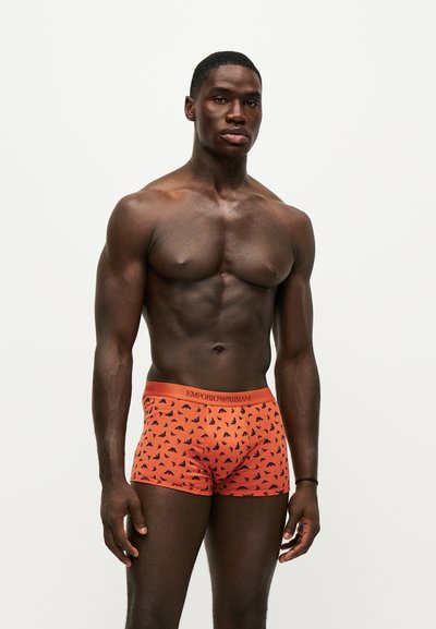 Homme musclé à la peau foncée debout, portant un boxer orange Emporio Armani avec un motif d'oiseau noir sur un fond blanc uni.