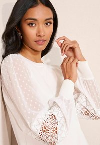 Fehér hosszú ujjú felső áttetsző pöttyös ujjakkal és csipkés szegéllyel a mandzsettánál, kerek nyakkivágással és puha kötött anyagból készült testtel.