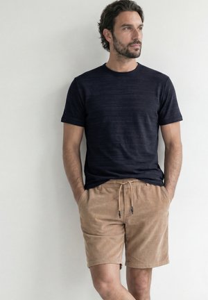 Mand stående med hænderne i lommerne iført sort kortærmet skjorte og beige snørede kordbukseshorts mod en hvid væg.