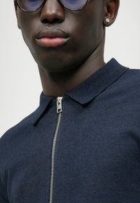 Polo de fecho éclair azul marinho feito de tecido texturizado, com colarinho e um detalhe de fecho éclair prateado. Óculos de sol redondos usados por cima.