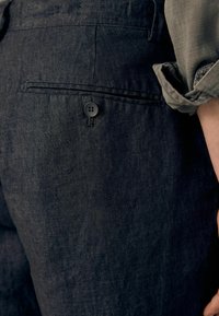 Pantalons en lin foncé avec une texture lisse, dotés d'une poche arrière unique et d'une fermeture à bouton noir. Manches retroussées d'une chemise olive visibles.