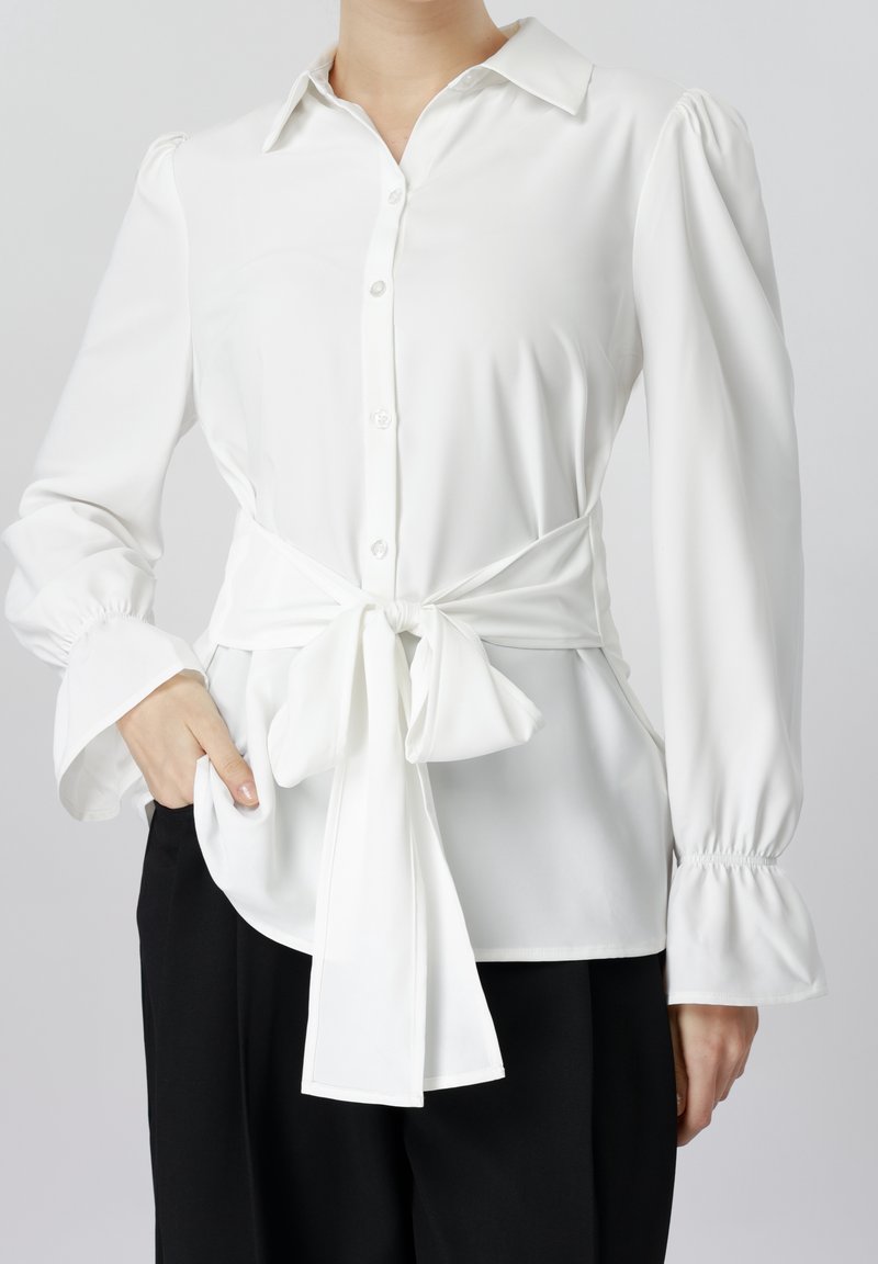 Femme portant un chemisier blanc à manches longues boutonné avec des manches bouffantes et un grand nœud noué à la taille, associé à un pantalon noir.