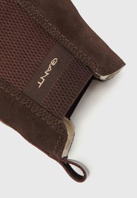 Close-up da parte de trás de uma bota de camurça castanha com painel elástico texturizado e aba de puxar, apresentando um forro de lã bege e um logotipo bege no elástico.