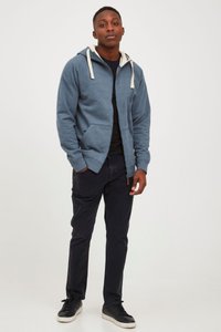 Blå zip-up hettegenser med kremfargede snører, brukt over en svart skjorte. Kombinert med mørke bukser og svarte joggesko. Har sidelommer.