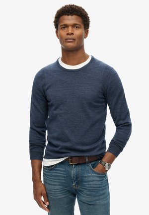 Suéter de punto azul marino con cuello redondo y mangas largas, llevado sobre una camisa blanca de cuello redondo, combinado con jeans azules y un reloj.