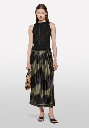 Top nero senza maniche con righe verticali, abbinato a una gonna di raso a patter verde e nero. La modella tiene una borsa intrecciata, indossando eleganti tacchi neri.