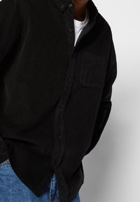 Camicia a bottoni in corduroy nera con tasca sul petto, caratterizzata da una superficie testurizzata e colletto classico, abbinata a jeans in denim blu.