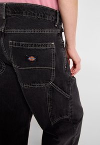 Black denim pants with contrasting white stitching, featuring a large rear pocket and a branded label. The fabric appears durable and textured.