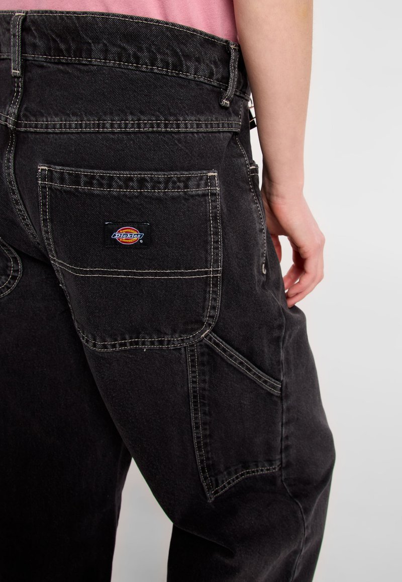 Pantaloni in denim nero con cuciture bianche a contrasto, dotati di una grande tasca posteriore e un'etichetta con il marchio. Il tessuto appare resistente e strutturato.