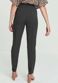 Pantalons noirs ajustés avec une coupe slim, présentant une texture lisse, des détails de plis subtils et des poches arrière. Associés à des chaussures à talons marron.