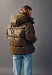 Brun puffjakke med hætte, med et quiltet design og blank overflade, parret med lyseblå jeans. Bagfra.