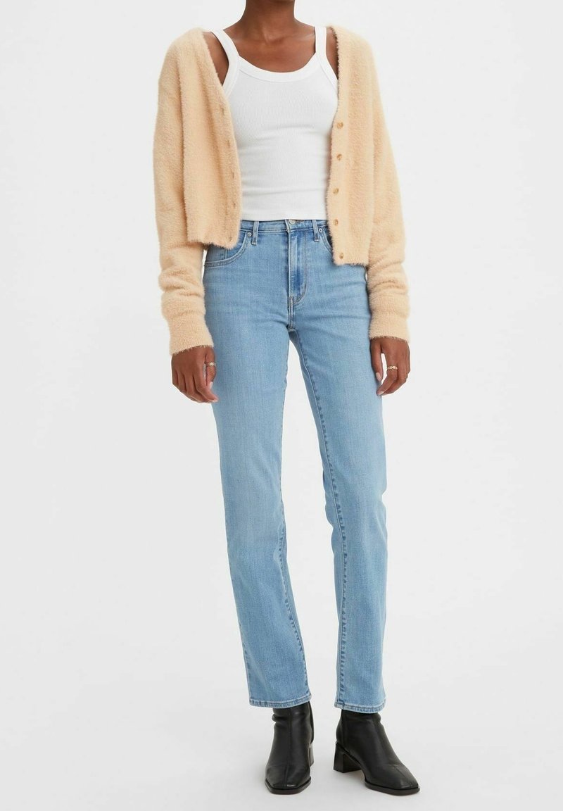 Helle beige, flauschige Strickjacke über einem weißen Tanktop, kombiniert mit hellblauen Jeans und schwarzen Knöchelstiefeln. Die Strickjacke hat ein Knopfdesign und lange Ärmel.