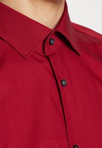 Rotes Hemd mit Knopfleiste, glatter Textur, klassischem Kragen und schwarzen Knöpfen, mit klaren Linien und einem taillierten Schnitt.