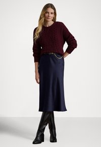 Bordó kábelmintás pulóver, sötétkék selyem midi szoknyával párosítva. Fekete térdig érő magassarkú csizma és aranylánc díszítés a övön.