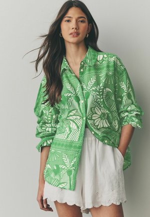 Cămașă verde cu guler butonat, prezentând un model floral și de frunze, cu accente albe. Cămașa are mâneci suflecate și o croială lejeră. Asortată cu pantaloni scurți albi cu margini ondulate.