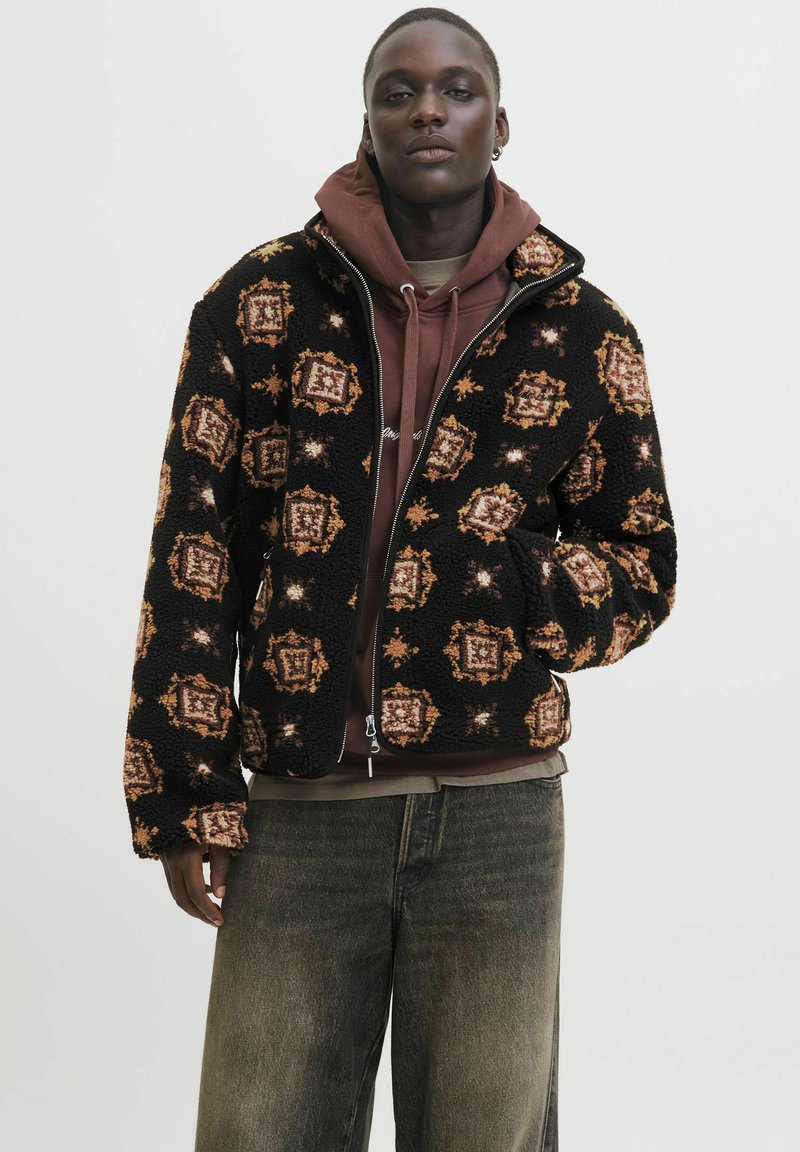 Giacca zip in fleece nera, con un motivo geometrico arancione e marrone. Indossata sopra un felpa marrone, con una vestibilità rilassata e un tessuto testurizzato.