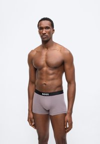 Μυώδης άνδρας όρθιος, φοράει γκρι μποξεράκια με μαύρη ζώνη μέσης στην οποία αναγράφεται "BOSS", πάνω σε απλό λευκό φόντο.