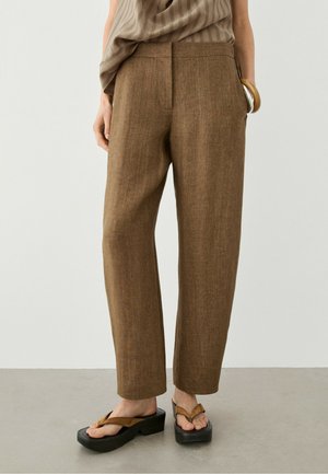 Femme portant un pantalon en lin marron à jambes droites, un haut marron sans manches, un bracelet en bois et des sandales à plateforme, debout sur un sol gris.