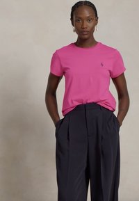 Rosa Baumwoll-T-Shirt mit kurzen Ärmeln und kleinem Logo, kombiniert mit marineblauen, hochgeschnittenen, gefalteten Hosen. Schlichtes, modernes Design.
