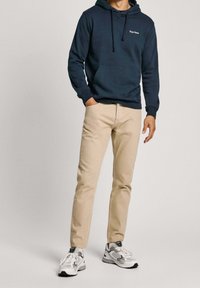 Felpa blu navy con cordini e tasca frontale, abbinata a pantaloni beige e sneakers grigie con dettagli neri. Outfit semplice e casual.