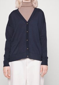 Marinblå cardigan med V-ringning, knäppning framtill och två framfickor. Mjuk stickad tyg med ribbade ärmslut och nederkant.