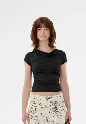 Ung kvinde med bølget brunt hår iført en tætsiddende sort top med korte ærmer og hvide bukser med sorte abstrakte pletter, stående mod en ensfarvet hvid baggrund.