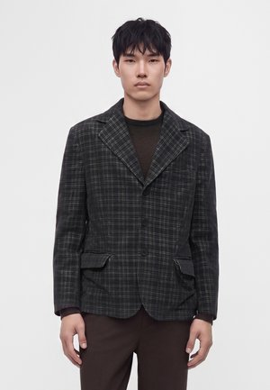 Homme aux cheveux courts noirs portant un blazer à carreaux gris foncé et un pantalon marron foncé, se tenant devant un fond clair uni.