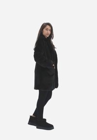 Sort faux fur frakke med høj krave, der har en afslappet pasform og siderlommer, parret med sorte leggings og ankelstøvler.