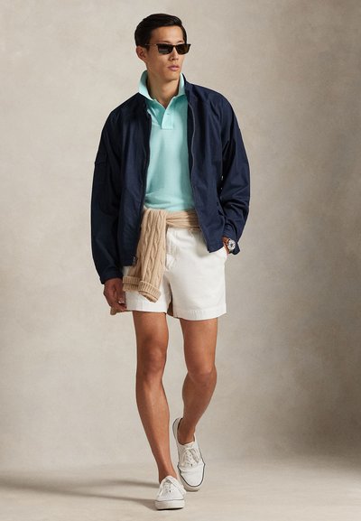 Μπλε ναυτικό μπουφάν, ανοιχτό μπλε πόλο, μπεζ πουλόβερ δεμένο στη μέση, λευκό σορτς, λευκά αθλητικά παπούτσια και σκούρα γυαλιά ηλίου. Λείο υλικό, casual στιλ.