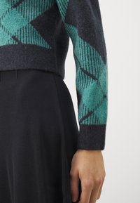 Strikkegenser med et geometrisk mønster i teal og svart, med en myk tekstur. Kombinert med et glatt svart stoff nedenfor.