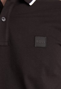 Dark brown polo shirt with a white striped collar. Features a textured black square logo patch with "BOSS" embossed. Smooth cotton material.