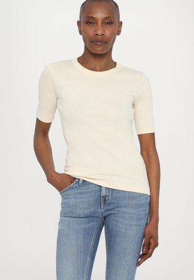 Camiseta ajustada de manga corta color crema con cuello redondo, hecha de una tela suave. Combinada con jeans de mezclilla azul claro.